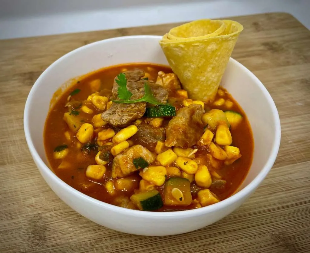 Beef caldo with corn and squash in a white bowl, a tortilla chip resting on top, on a wooden board.
