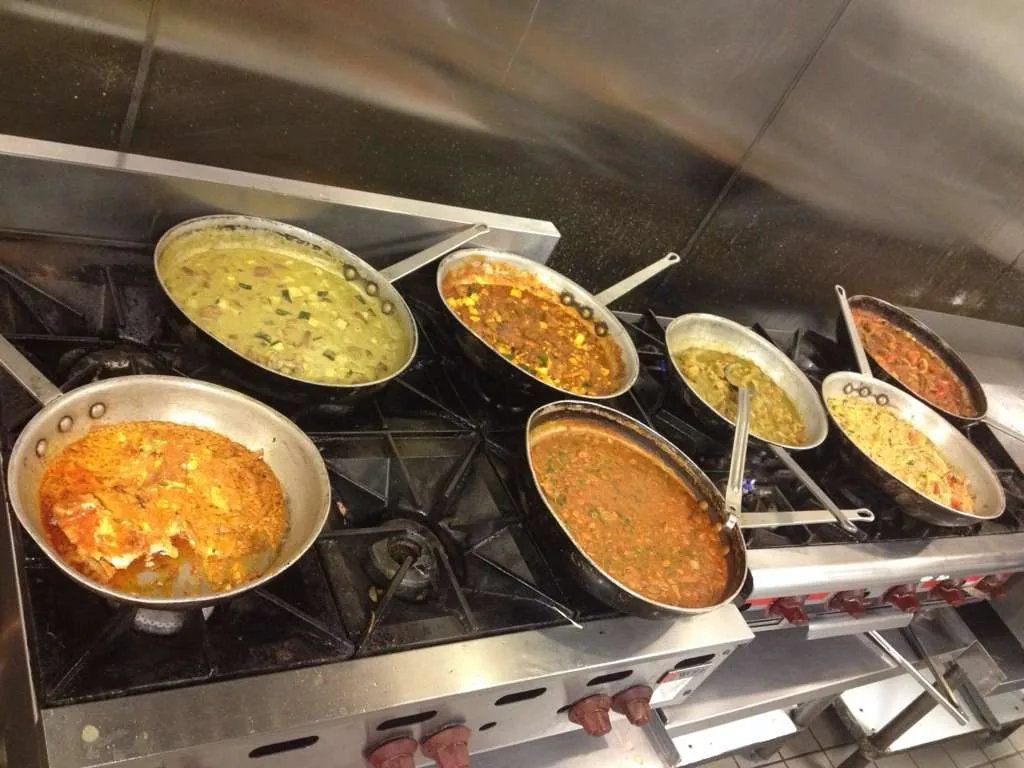 Seven pans of different guisados, all on the fire at the same time, on a steel industrial range.