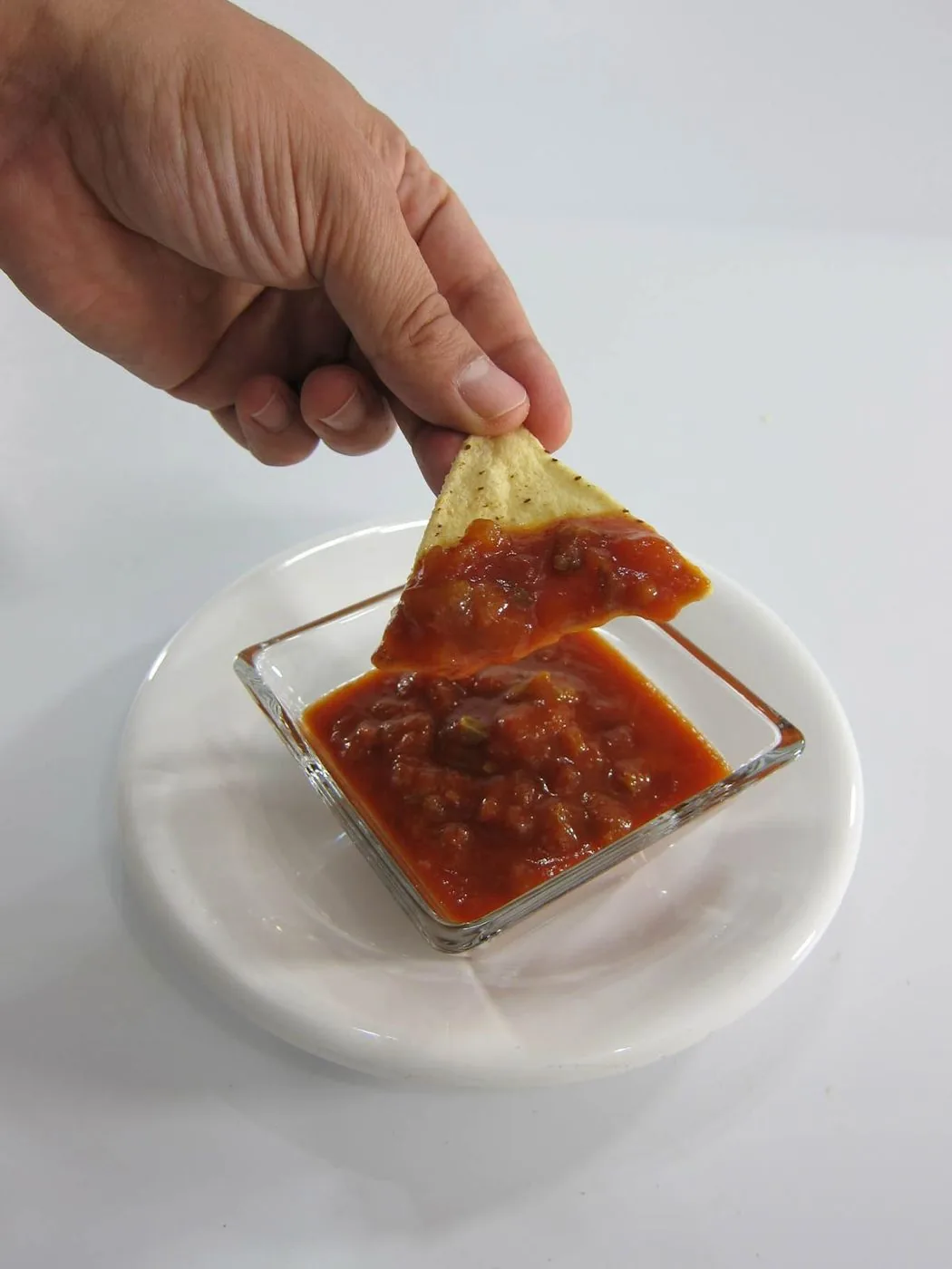 A hand dipping a tortilla chip into a square glass bowl of red salsa.