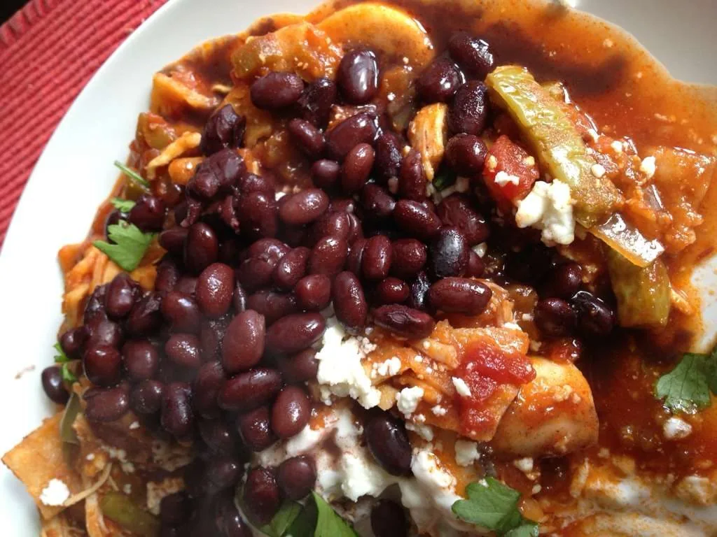 Black beans over a guisado with strips of chile, queso fresco and cilantro, on a white plate.