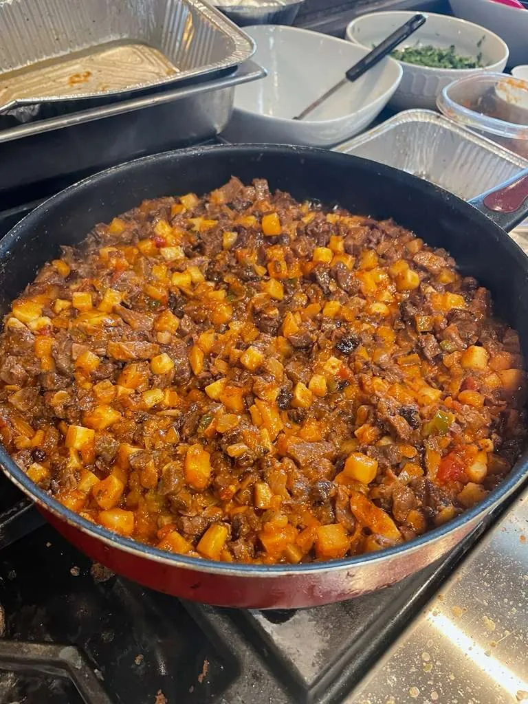A large pot of picadillo with potato on a stove, steel trays behind it.