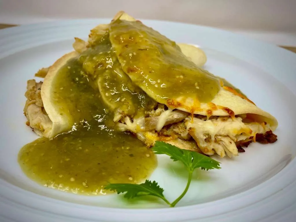 A chicken quesadilla on a white plate, under a pour of salsa verde with strips of chile.
