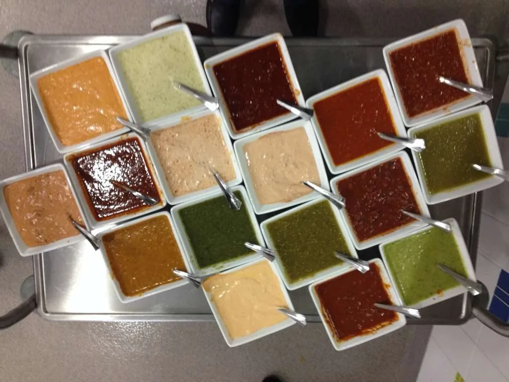 Overhead view of a steel tray holding more than a dozen different salsas in white containers, each one with its own spoon.