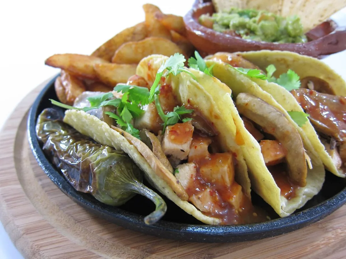 Tacos in a cast-iron skillet on a wooden board, with browned potatoes, guacamole and tortilla chips.