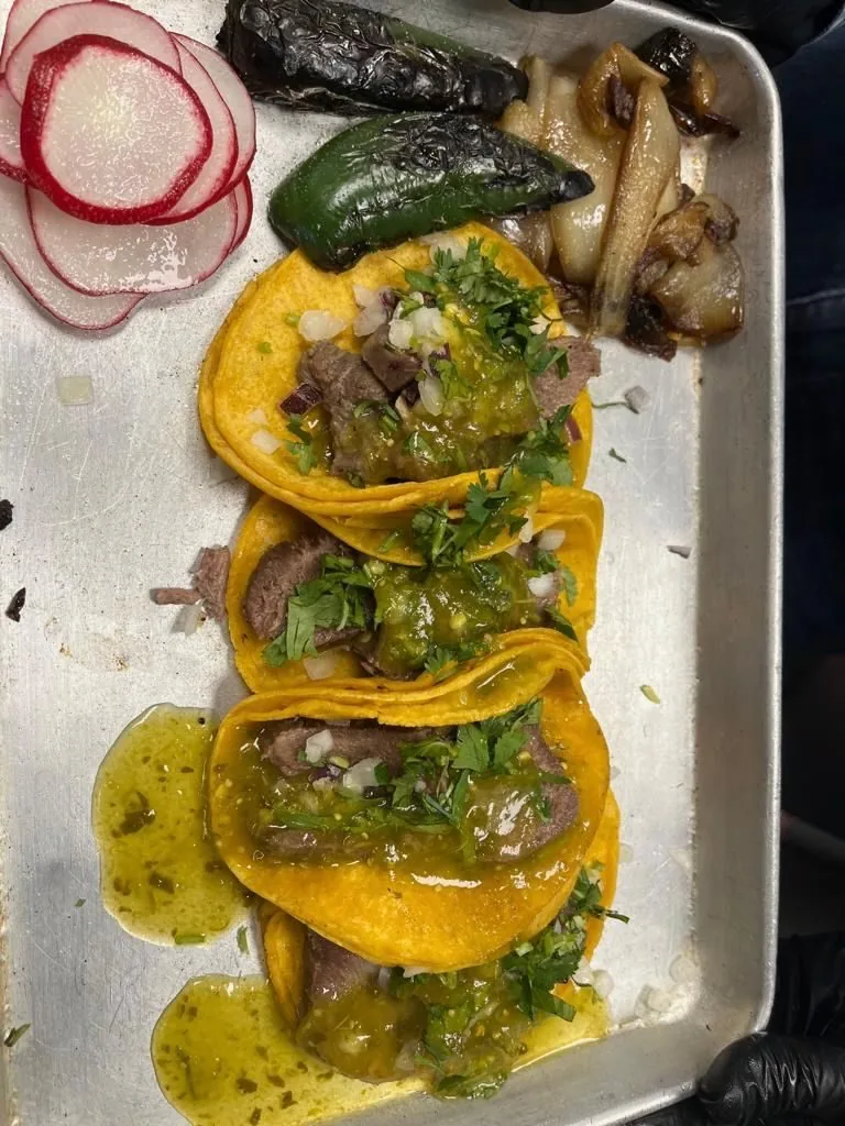 Three beef tacos with salsa verde and cilantro on a steel tray, with sliced radish, roasted chiles and onion.