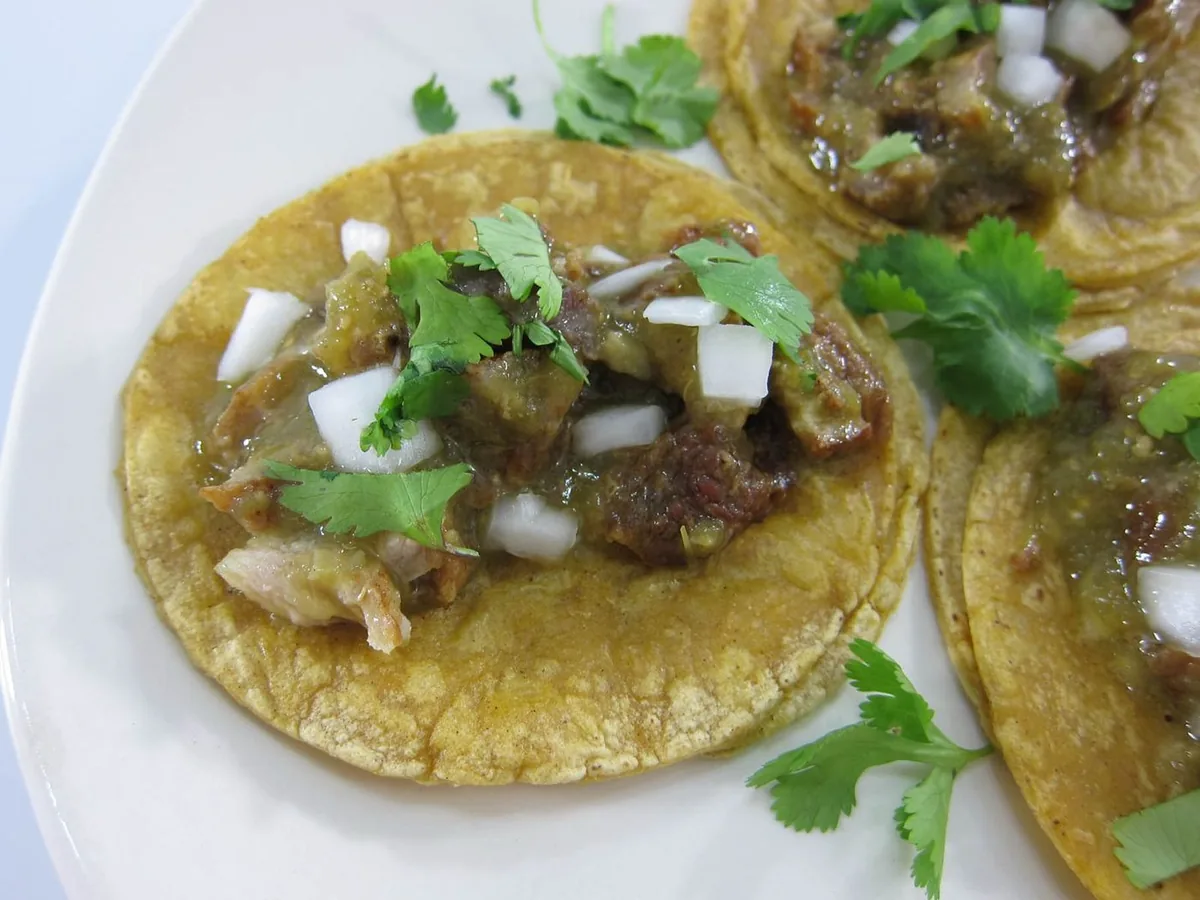 Corn tortilla tacos with meat, salsa verde, onion and cilantro on a white plate.
